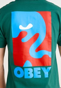 Tricou verde cu un grafic mare pe spate, având forme abstracte roșii și albastre și cuvântul "OBEY" în litere albastre îngroșate.