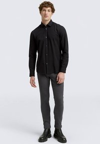 Juoda marškiniai su sagomis iš lengvos medžiagos, derinami su aptemptomis pilkomis kelnėmis ir aulinukais. Modelis stovi neutraliu pozu priešais šviesią foną.