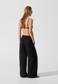 Pantalon noir à jambes larges avec une taille élastique, associé à un haut de bikini rayé foncé. Modèle de dos, cheveux mouillés et brillants, portant des lunettes de soleil.
