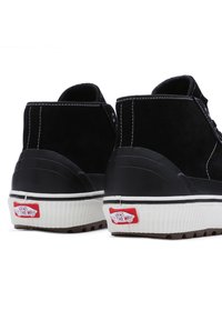 Baskets montantes en daim noir avec des semelles en caoutchouc blanc, des coutures contrastantes et un patch logo Vans rouge sur le talon.