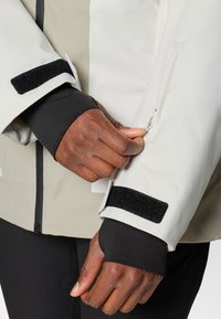 Vedenkestävä takki, jossa on kerma-valkoinen päällyskerros, mustat hihansuut ja tarrakiinnitys. Varustettu sivuvetoketjulla ja rakenteellisella istuvuudella.