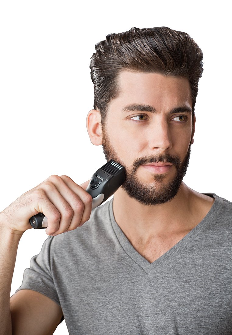 Un cortapelos negro y gris sostenido por un hombre con barba, vestido con una camiseta gris de cuello en V, concentrado en arreglar su vello facial.