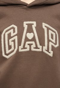 Καφέ ύφασμα με μεγάλες λευκές υφασμένες γράμματα που σχηματίζουν τη λέξη GAP, με ένα σχήμα καρδιάς στο κέντρο του γράμματος Α.