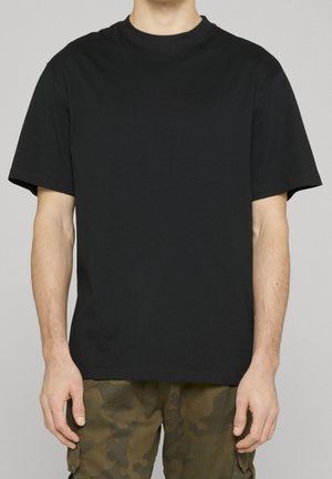 Person iført en ensfarvet sort kortærmet T-shirt og grøn camouflage cargo bukser, stående mod en ensfarvet baggrund.