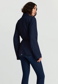 Giacca blu navy con vita aderente, design con cintura e maniche larghe. Presenta una spaccatura sul retro ed è abbinata a jeans scuri.