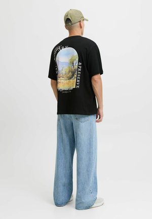 Mand iført sort t-shirt med farverigt naturmotiv og tekst, lys blå brede bukser, hvide sneakers og en grøn baseballkasket, der vender væk.