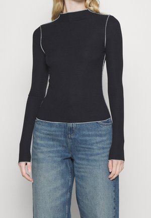 Femme portant un haut noir ajusté à manches longues avec des surpiqûres blanches contrastantes et un jean bleu taille haute sur un fond uni.