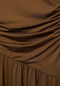 Hnědé šaty s hladkým, lehkým materiálem. Mají na vrchu nařasené detaily a rozšířený volánkový lem.