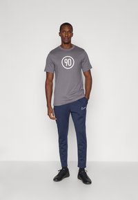 T-shirt in cotone grigio con una grafica circolare bianca che legge "90"; abbinato a pantaloni sportivi blu navy e scarpe nere. Design semplice.