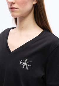 Γυναίκα που φοράει μαύρο πουκάμισο με V λαιμόκοψη και γκρίζο λογότυπο "CK" και λευκό "Calvin Klein" στο αριστερό στήθος, με ασημένιο κρίκο σκουλαρίκι ορατό.