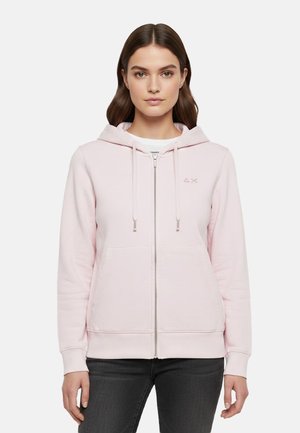 Donna che indossa una felpa con cappuccio rosa chiaro con cerniera, tasche frontali e coulisse, abbinata a jeans scuri, in piedi davanti a uno sfondo semplice.