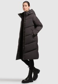 Long black puffer coat with a hood, quilted design, zipper closure, and side pockets, worn over black leggings and chunky black boots.