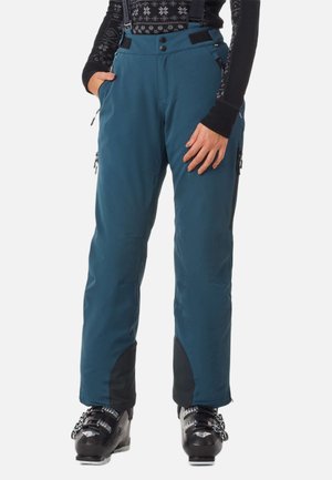 Blauwe skibroek met bretels, gedragen met zwarte skischchoenen en een zwart-patroon langemouwen top, handen rustend bij de zakken.