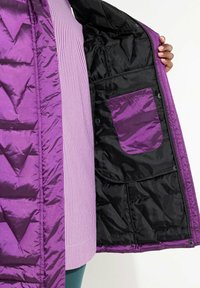 Lila pufferjacka med geometrisk quiltning, som har en svart insida och en liten ytterficka. Tyget har en glansig yta.
