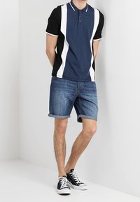 Polo shirt blu navy e nera con strisce bianche, colletto a due bottoni, abbinato a pantaloni di jeans corti arrotolati e sneakers nere. Vestibilità casual.