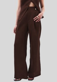 Hnědé široké kalhoty vyrobené z lehkého materiálu, s plisovaným designem a bočními kapsami. Nosí se s černými sandály a minimálními doplňky.