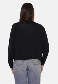 Cardigan nero corto con maniche lunghe. Dettaglio a costine sull'orlo e texture morbida. Modello indossa jeans azzurri chiari. Vista da dietro.