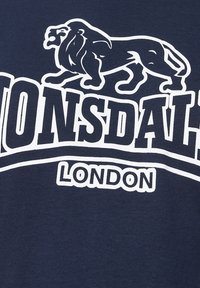 Tengerészkék póló fehér oroszlán grafikával és "LONSDALE LONDON" szöveggel, vastag, blokk betűkkel. Simogatós pamut anyag, laza dizájnnal.
