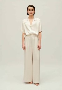 Bēšas, brīvi pieguļošas drēbes, kas sastāv no īsām piedurknēm ar apkakli un platām biksēm ar elastīgu jostu, fona ir neitrāls.