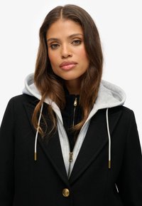 Zwarte wollen jas met gouden knoop, met daaronder een lichtgrijze hoodie. Soepele textuur met een gestructureerd silhouet.