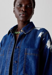 Giacca in denim blu scuro con macchie di vernice bianca, chiusura a bottoni, due tasche frontali e dettagli di cucitura a contrasto.