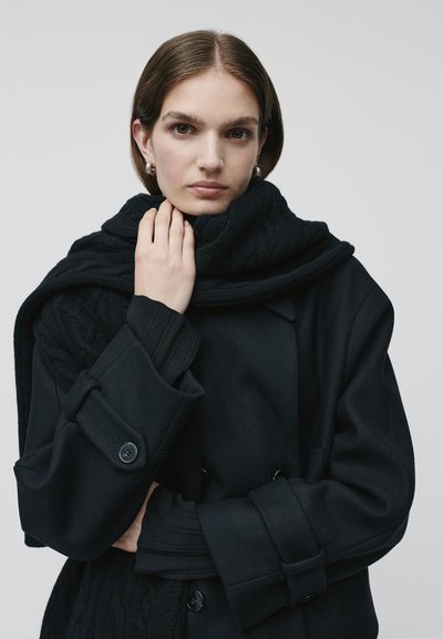 Cappotto doppiopetto nero con sciarpa in maglia a treccia spessa; entrambi gli articoli sono caratterizzati da tessuti testurizzati di diversi tipi e presentano cuciture dettagliate.