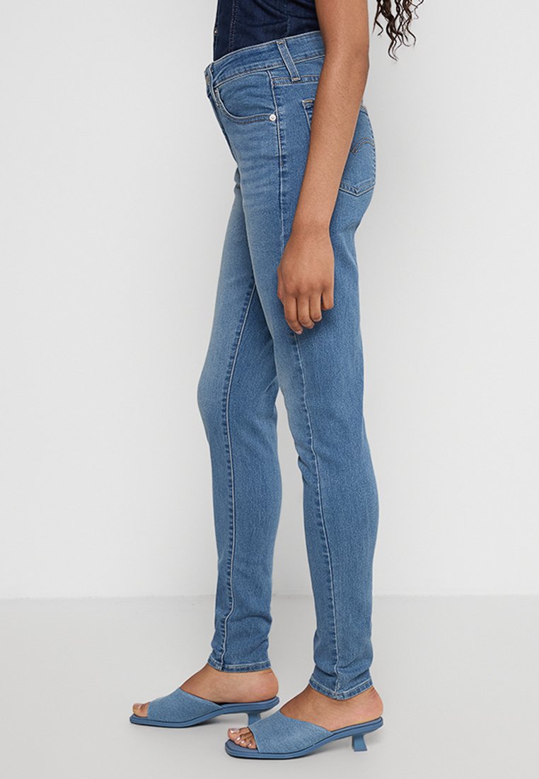 Jeans en denim bleu taille haute avec une coupe slim, comprenant une poche avant, des coutures ton sur ton, et associés à des talons ouverts bleu clair.