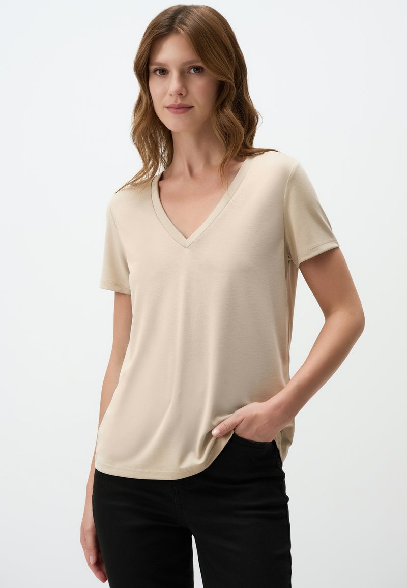 T-shirt beige a scollo a V in morbido tessuto, maniche corte, vestibilità rilassata, con dettagli di cucitura discreti, abbinata a pantaloni neri.