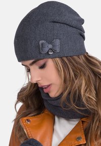 Woman wearing a gray knit hat with a bow and matching neck warmer, paired with an orange leather jacket and white top.
