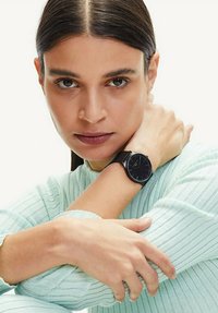 Zwarte analoge horloge met een gestructureerde mesh-band, met een eenvoudige ronde wijzerplaat en minimalistisch ontwerp. De achtergrond is licht.