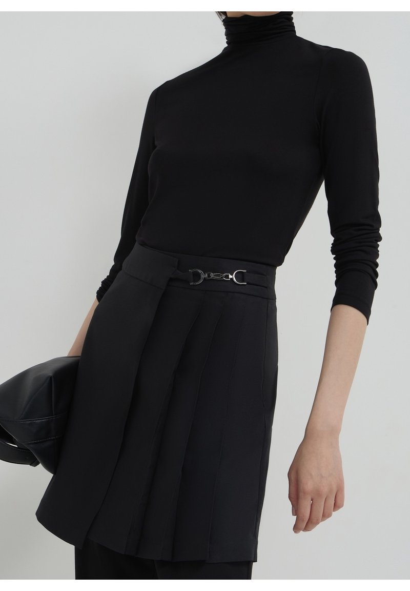 Personne portant un col roulé noir et une jupe noire taille haute avec une ceinture à boucle décorative, tenant un sac noir dans la main gauche.