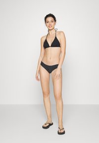 Biquíni preto com um top triangular em tecido texturizado e parte de baixo de cintura alta com riscas. Modelo complementou o visual com chinelos.