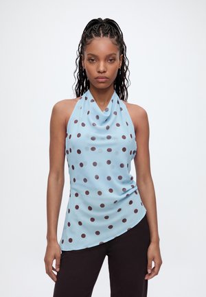 Femme aux cheveux tressés portant un haut dos nu bleu clair à pois foncés et un pantalon noir sur un fond blanc uni.