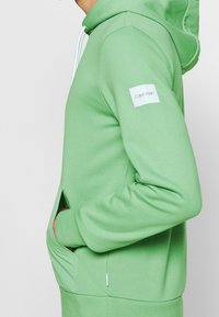 Felpa verde con una vestibilità rilassata, realizzata in tessuto morbido. Presenta una tasca frontale, cappuccio con coulisse e un logo bianco di Calvin Klein sulla manica.