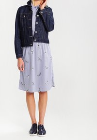 Mörk denimjacka över en randig klänning med fjärilsbroderi, kombinerad med mörka sneakers. Rakt snitt, korta ärmar, knälång klänning.