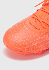 Klar orange fodboldstøvle med tekstureret strikket overdel, der har reflekterende accenter og standard snøresystem; klare plastikstifter under.