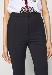 Pantaloni neri aderenti con una texture liscia, vita alta e dettagli minimi, mostrati dalla vita in giù con le mani ai lati.