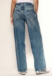 Jeans de mezclilla azul de pierna ancha con un acabado desgastado, que presentan dos bolsillos traseros y un ajuste relajado, combinados con bailarinas plateadas.