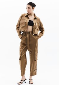 Geknipte bruine jas met twee voorzakken, gecombineerd met bijpassende cargo-broek met riem en zijzakken, met boorden. Zwarte top en sandalen.