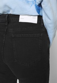 Mustat farkut, joissa on vartalonmyötäinen muotoilu, kaksi takataskua, ommeltu yksityiskohtia ja vaatelappu osittain näkyvissä vyötäröllä.