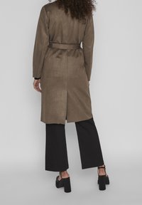 Manteau long marron avec une ceinture à la taille, présentant une texture lisse et une fente à l'arrière, associé à un pantalon large noir et des sandales à talons.