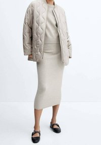 Quilted beige bomberjakke med rund krave, parret med en tætsiddende beige strikkjole og sorte flade sko med dekorative stropper.