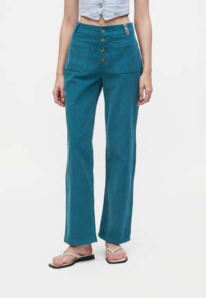 Femme portant un pantalon en velours côtelé sarcelle taille haute avec des boutons devant et des poches plaquées, assorti à des sandales beiges et un top court bleu clair.
