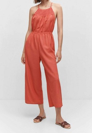 Femme portant une combinaison corail sans manches avec des jambes larges et des sandales plates marron, debout devant un fond clair uni.