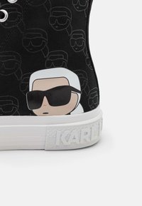 Baskets noires à semelle blanche présentant des visages de dessins animés répétés avec des lunettes de soleil et le nom "KARL" embossé sur la semelle.