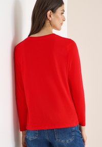 Rotes Langarmshirt aus glattem Stoff, mit rundem Ausschnitt und leicht lockerer Passform, kombiniert mit blauen Jeans.