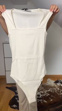 Mãos seguram um bodysuit branco sem mangas com decote quadrado contra um fundo neutro interior com piso de madeira e tapete.