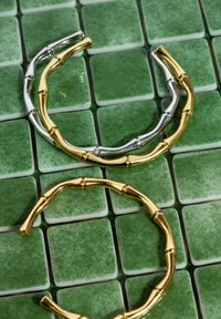 Due bracciali a forma di bambù in tonalità argento e oro, posizionati su piastrelle quadrate smaltate di verde, evidenziando finiture metalliche lucide e una texture liscia.