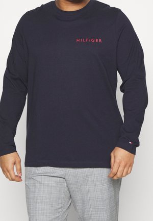 Mężczyzna ubrany w granatową koszulę z długim rękawem z czerwonym napisem "HILFIGER" oraz jasnoszare spodnie w kratę, stojący na jednolitym tle.