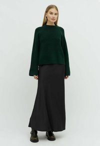 Pulover verde tricotat cu guler rotund și mâneci largi, asortat cu o fustă maxi neagră, cizme până la glezne și texturi fine.